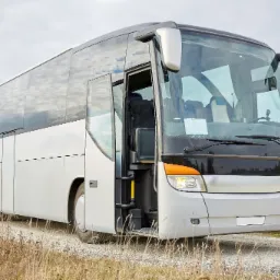 Nowoczesny autobus turystyczny z otwartymi drzwiami wejściowymi, zaparkowany na żwirowej drodze z widoczną suchą trawą.