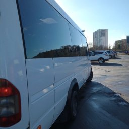 Busowicz spółka z ograniczoną odpowiedzialnością - Biały bus pasażerski zaparkowany na parkingu, z odbiciem budynków w przyciemnianych szybach.