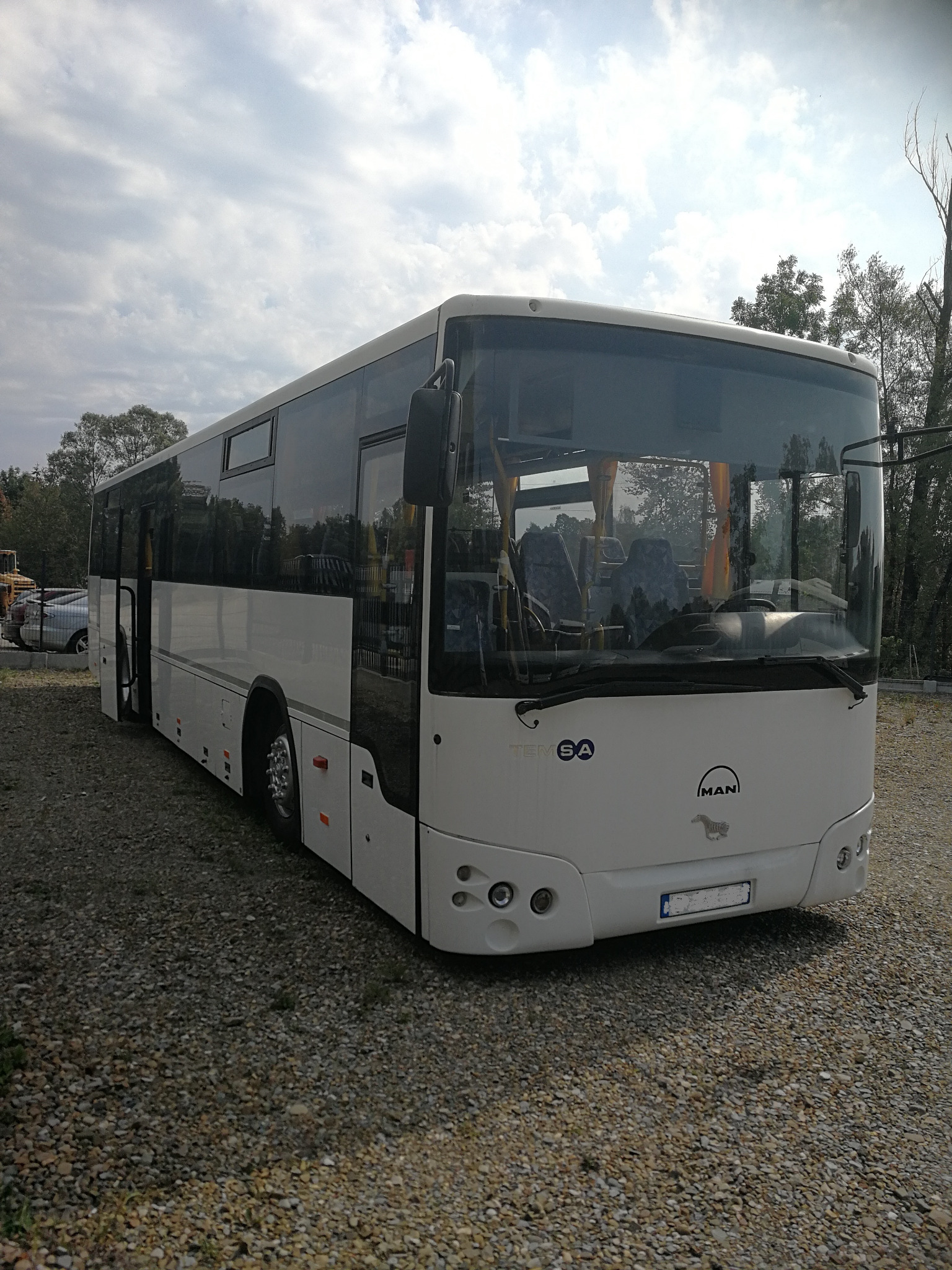 Biały autobus marki MAN firmy z Poznania, zaparkowany na żwirowym terenie pod pochmurnym niebem.