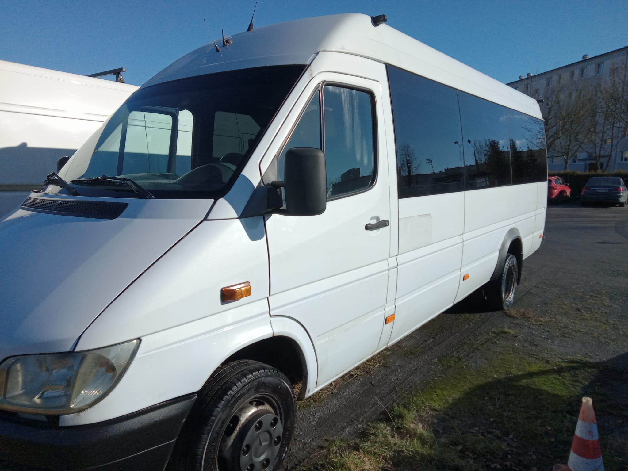 Biały bus pasażerski Mercedes-Benz Sprinter z ciemnymi szybami, zaparkowany na żwirowym podłożu, widoczny stożek ostrzegawczy.