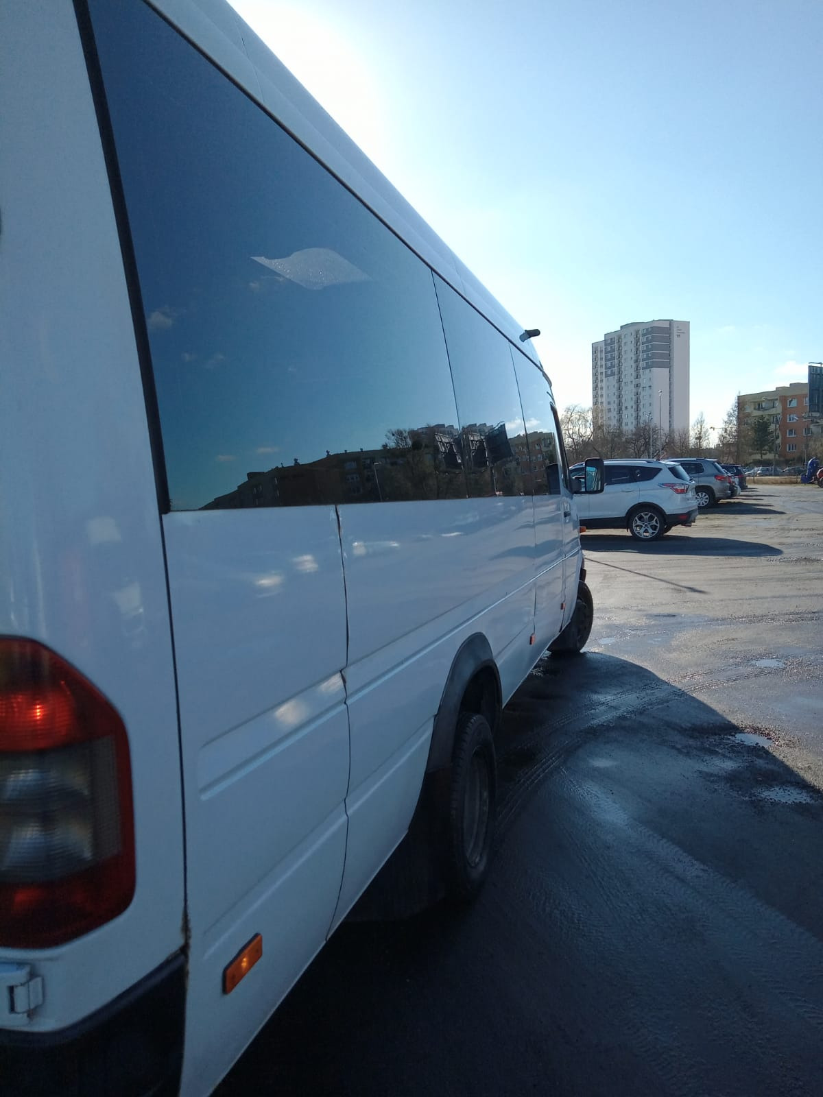 Biały bus pasażerski zaparkowany na parkingu, z odbiciem budynków w przyciemnianych szybach.