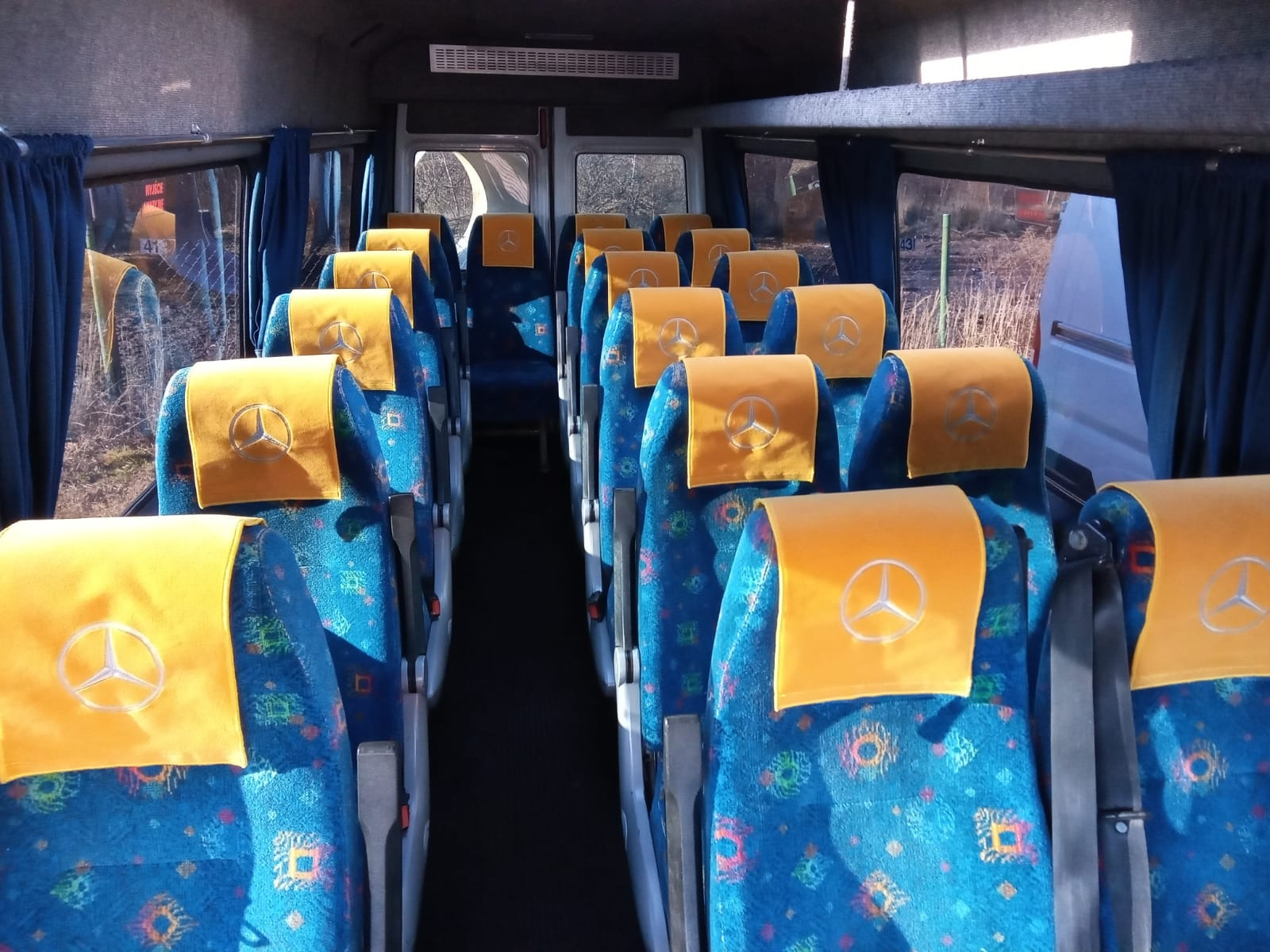 Wnętrze autobusu z niebieskimi fotelami w kolorowy wzór i żółtymi zagłówkami z logo Mercedes-Benz, widok z przodu na puste siedzenia.