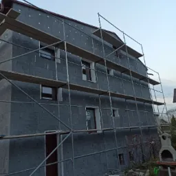 Dom w trakcie nakładania warstwy izolacyjnej na elewacji, widoczne rusztowania i przygotowane otwory okienne i drzwiowe, na podwórku pozostałości materiałów budowlanych.