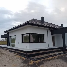 Nowoczesny, parterowy dom z białą elewacją i ciemną stolarką okienną, widoczny z zewnątrz, z elementami wykończeniowymi w trakcie realizacji.