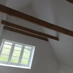 Poddasze z oknem dachowym i widocznymi belkami stropowymi, świeżo pomalowane na biało, z wystającymi kablami elektrycznymi.