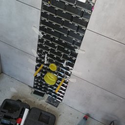 Usługi remontowe - Montaż błyszczących, czarnych płytek typu metro w pionowym pasie na szarej ścianie, z użyciem krzyżyków dystansowych i taśmy maskującej; widoczne narzędzia, poziomica i butelka wody.