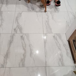 Usługi remontowe - Białe, marmurowe płytki podłogowe ułożone w kwadraty w Włostowie, widoczne nogi kobiety w czarnych klapkach z pomponami i pies rasy pitbull.