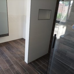 Usługi remontowe - Nowoczesna łazienka z brązowymi płytkami imitującymi drewno na podłodze i ścianach prysznica, białe płytki z wnęką w ścianie, szklana kabina prysznicowa z czarnymi elementami wykończeniowymi.