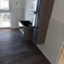 Usługi remontowe - Wnętrze kuchni w trakcie montażu: ciemne płytki podłogowe imitujące drewno, białe płytki ścienne z delikatnym wzorem, częściowo zamontowana szafka pod zlew z lustrzanymi frontami, okno z widokiem...