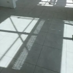 Ułożone szare płytki podłogowe w jasnym pomieszczeniu, z wyraźnym cieniem rzucanym przez okno, widoczne instalacje przy ścianie i fragment drewnianej podłogi przy krawędzi kadru.