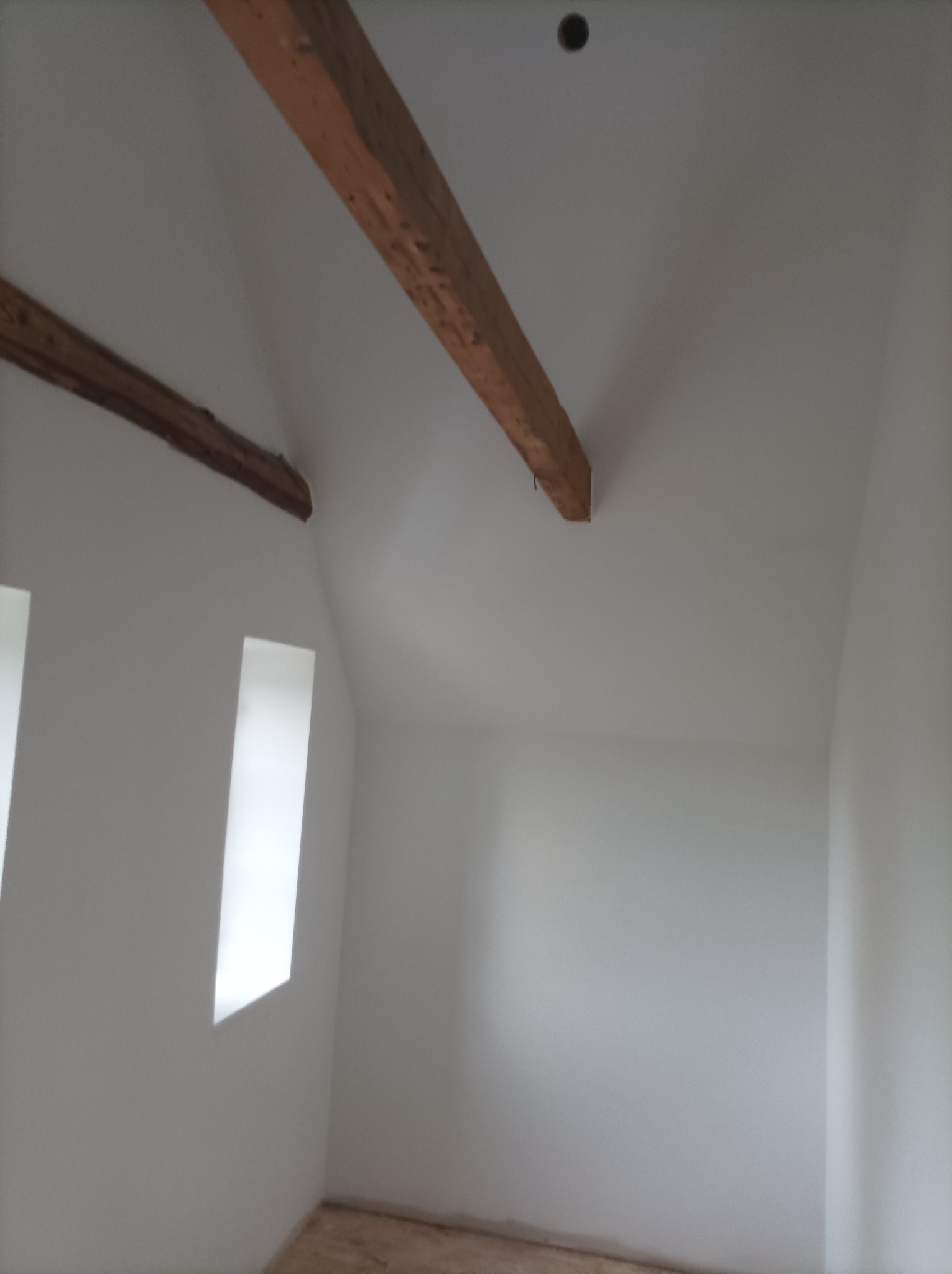 Poddasze po remoncie: białe ściany, drewniane belki stropowe, dwa okna w skosach, podłoga częściowo widoczna.