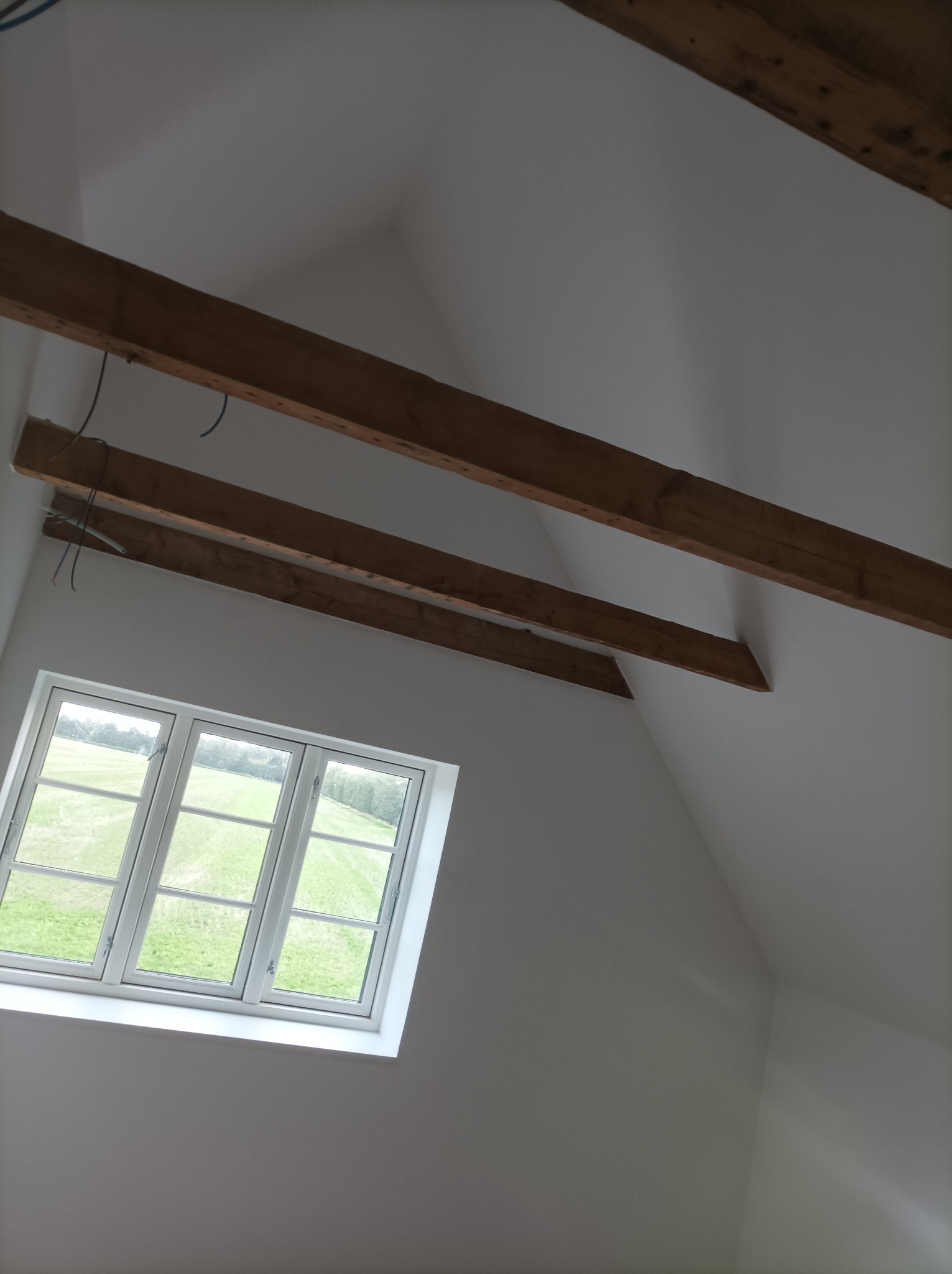 Poddasze z oknem dachowym i widocznymi belkami stropowymi, świeżo pomalowane na biało, z wystającymi kablami elektrycznymi.