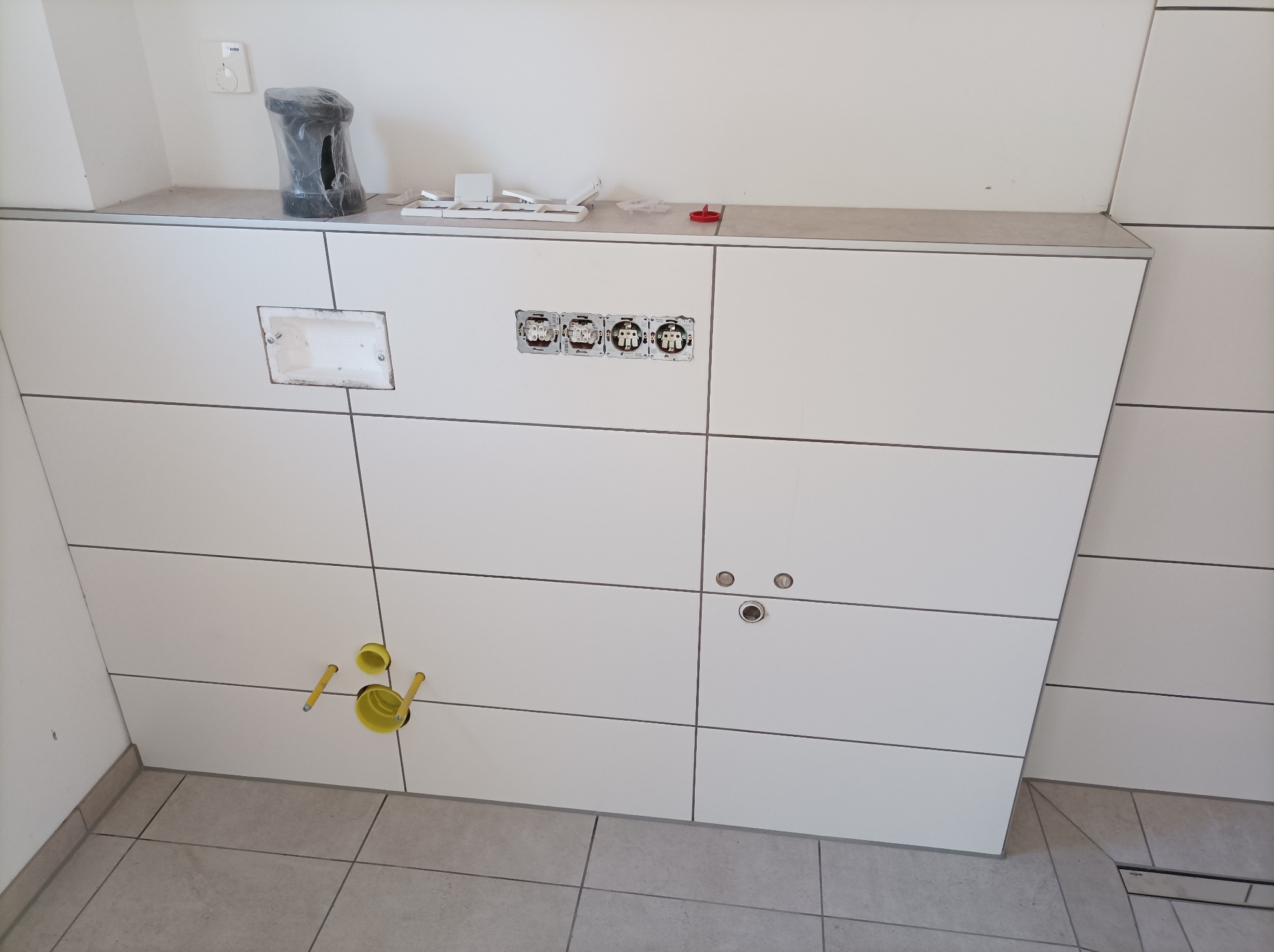 Ściana w łazience wyłożona białymi, prostokątnymi płytkami z widocznymi otworami na gniazdka elektryczne, odpływ wody i podłączenia kanalizacyjne z żółtymi rurami.