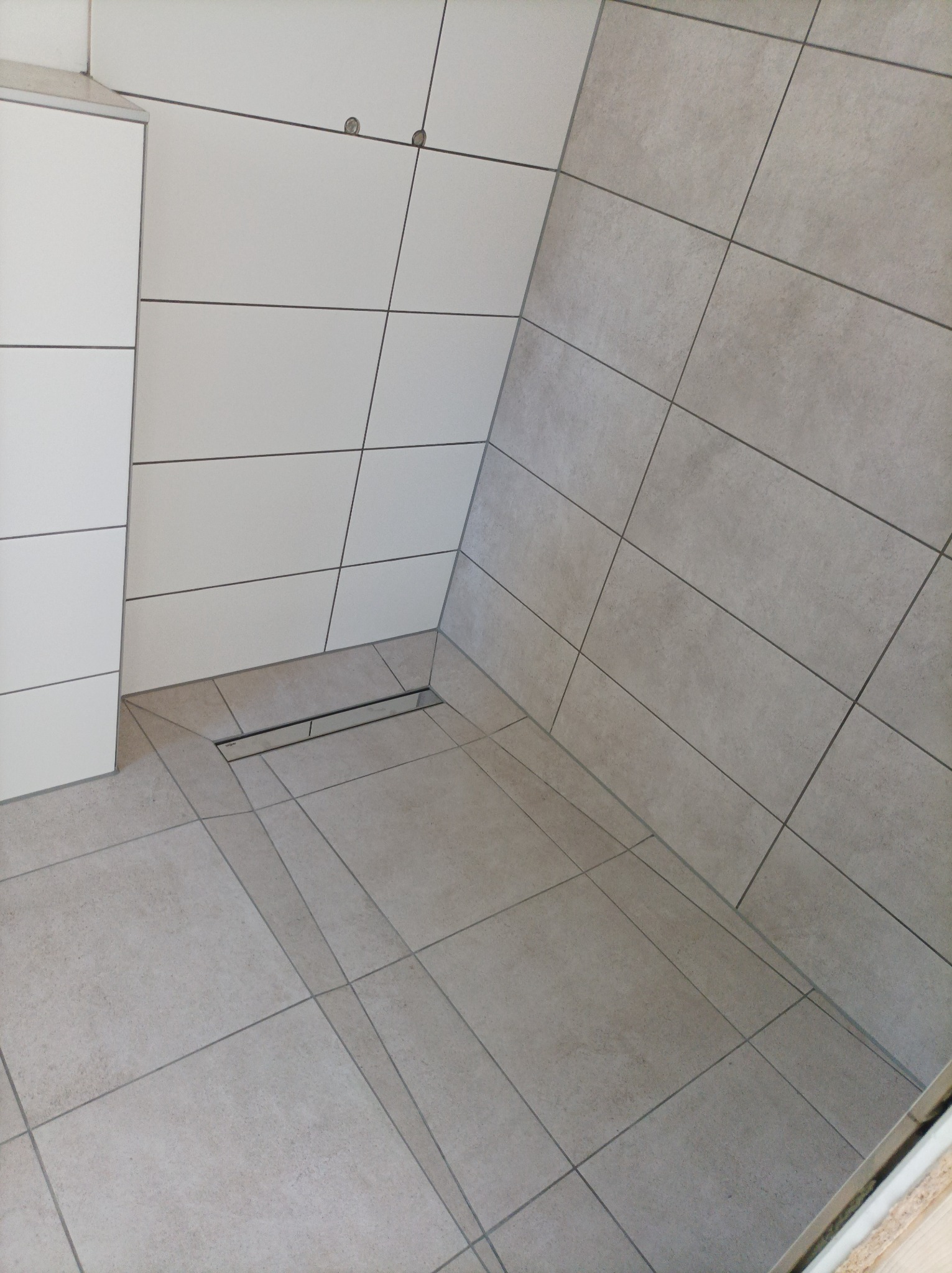 Wykończona łazienka w odcieniach szarości z odpływem liniowym w podłodze i zabudową meblową w rogu, płytki ułożone pod skosem.