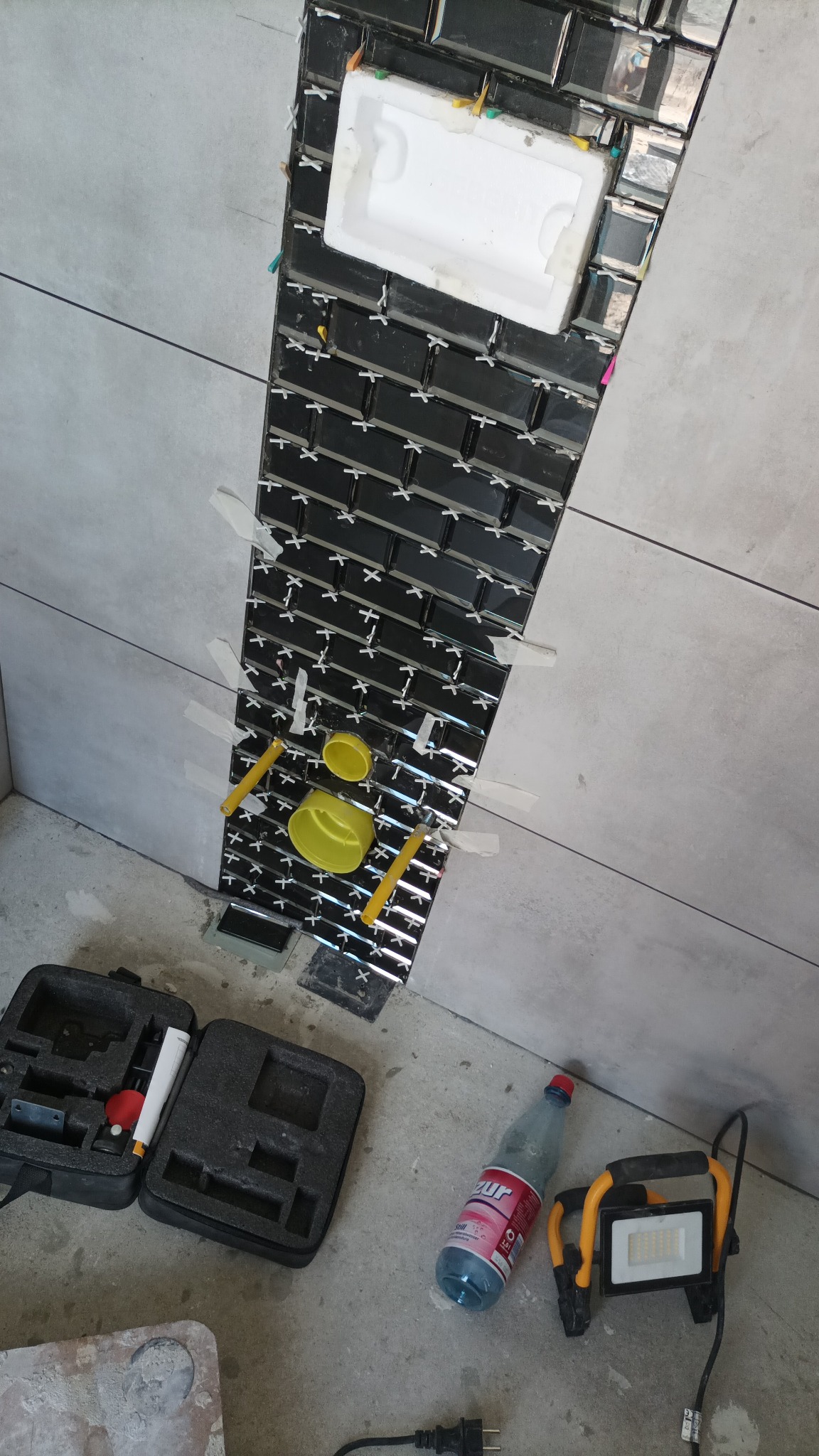 Montaż błyszczących, czarnych płytek typu metro w pionowym pasie na szarej ścianie, z użyciem krzyżyków dystansowych i taśmy maskującej; widoczne narzędzia, poziomica i butelka wody.