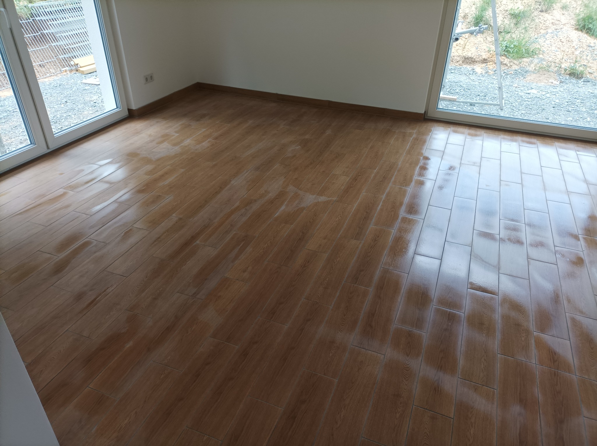 Ułożona, błyszcząca podłoga z płytek imitujących drewno w pustym pokoju z dużymi oknami balkonowymi, widoczne ślady po czyszczeniu na powierzchni.