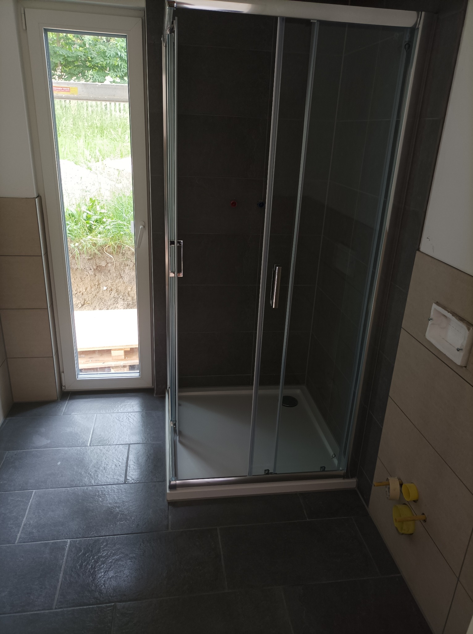 Nowoczesna kabina prysznicowa z przesuwnymi drzwiami w łazience z ciemną, kamienną podłogą i jasnymi, beżowymi płytkami ściennymi; widoczne wystające rury z żółtymi zaślepkami i okno z widokiem...