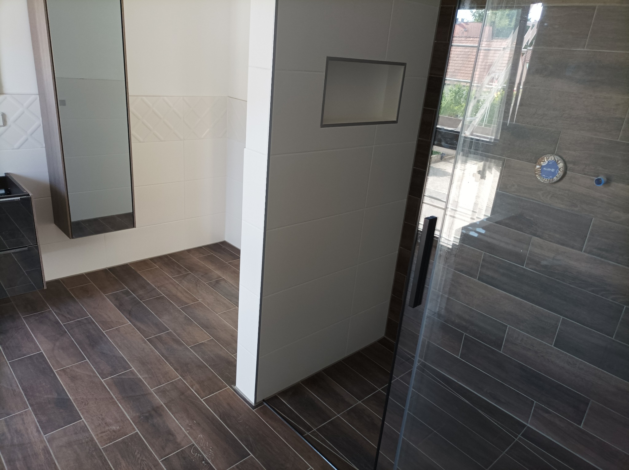 Nowoczesna łazienka z brązowymi płytkami imitującymi drewno na podłodze i ścianach prysznica, białe płytki z wnęką w ścianie, szklana kabina prysznicowa z czarnymi elementami wykończeniowymi.