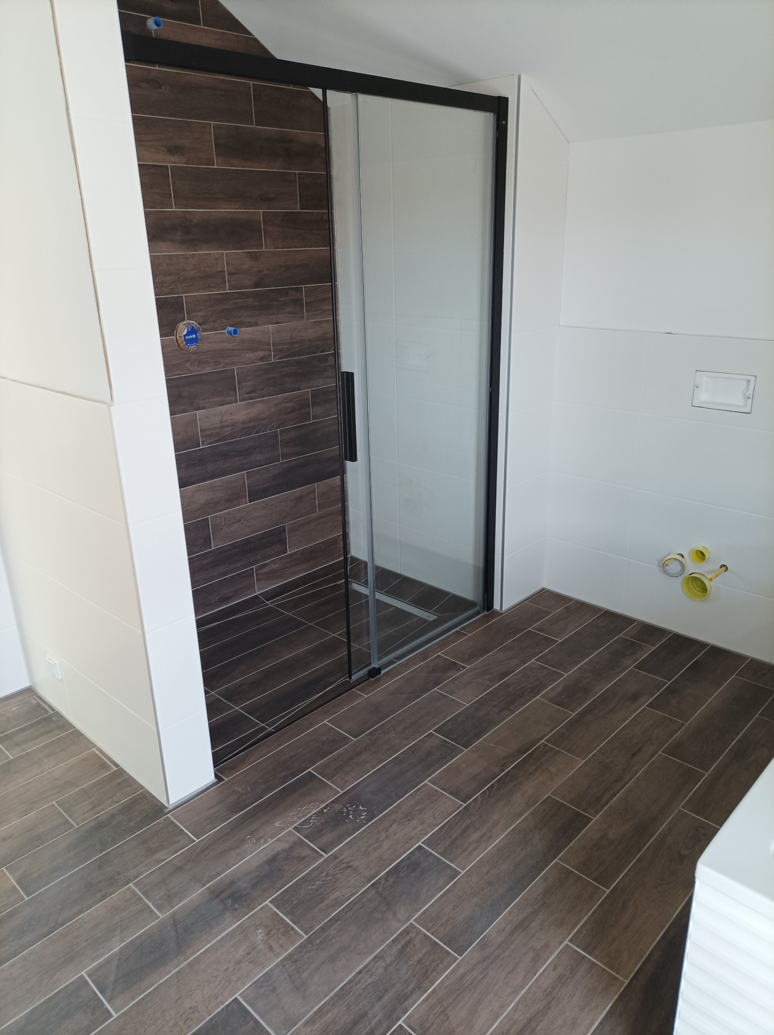 Nowoczesna łazienka z kabiną prysznicową typu walk-in, wykończona płytkami imitującymi drewno w odcieniach brązu, z widocznymi przyłączami hydraulicznymi przed montażem armatury.