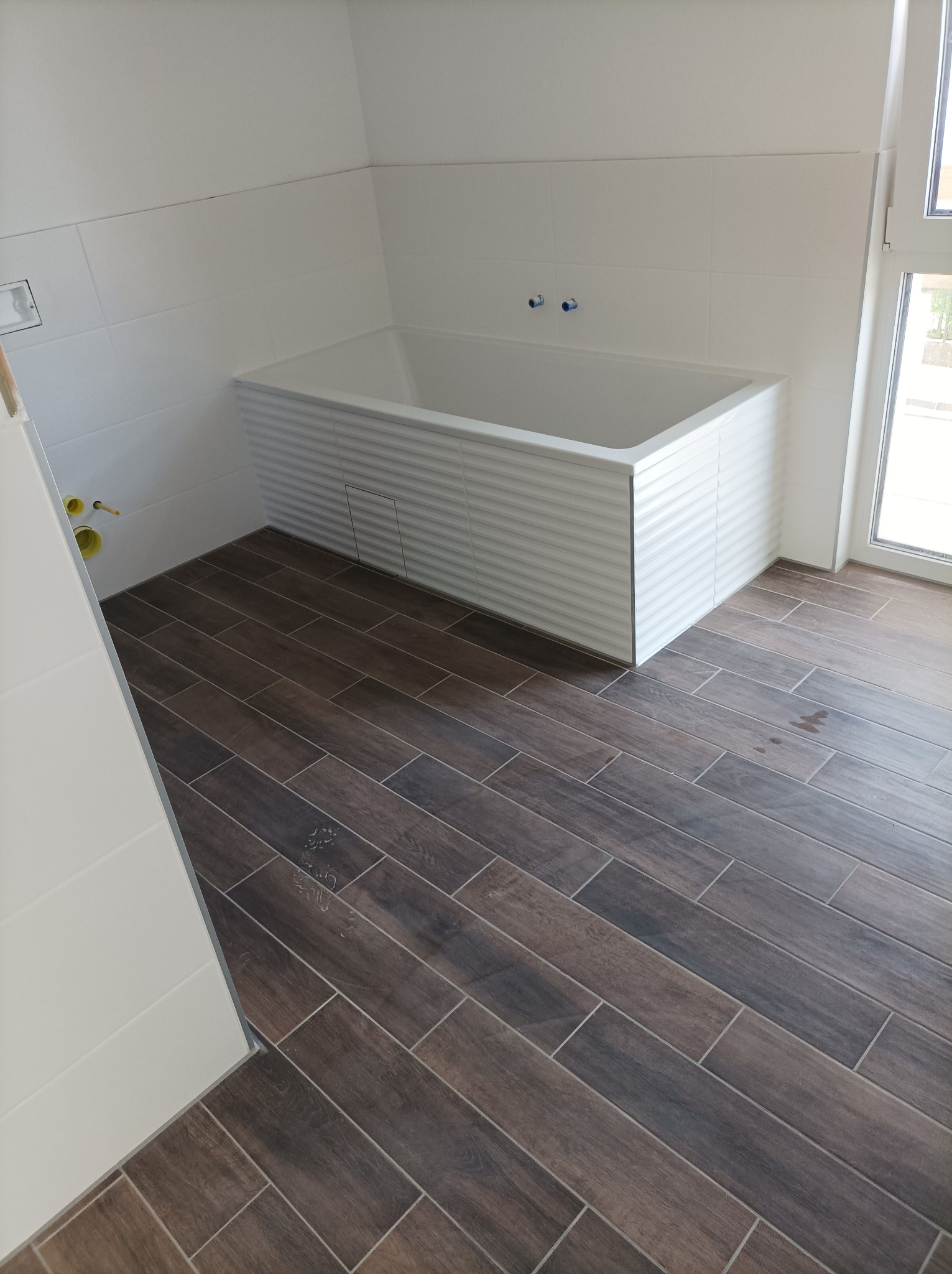 Nowoczesna łazienka z wanną obudowaną białymi panelami i podłogą wyłożoną ciemnobrązowymi płytkami imitującymi drewno, widoczne przyłącza wodne i fragment okna.