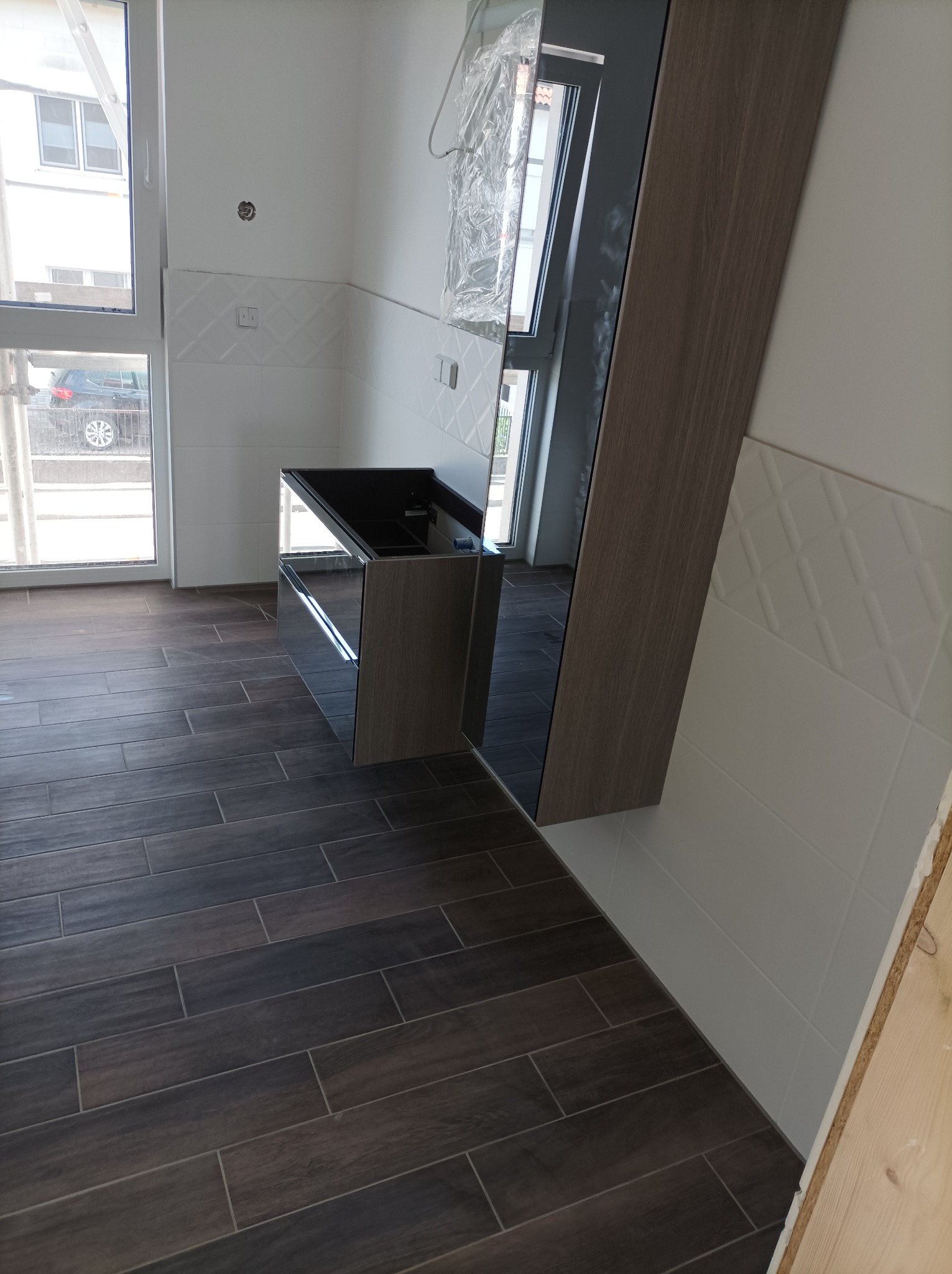 Wnętrze kuchni w trakcie montażu: ciemne płytki podłogowe imitujące drewno, białe płytki ścienne z delikatnym wzorem, częściowo zamontowana szafka pod zlew z lustrzanymi frontami, okno z widokiem...