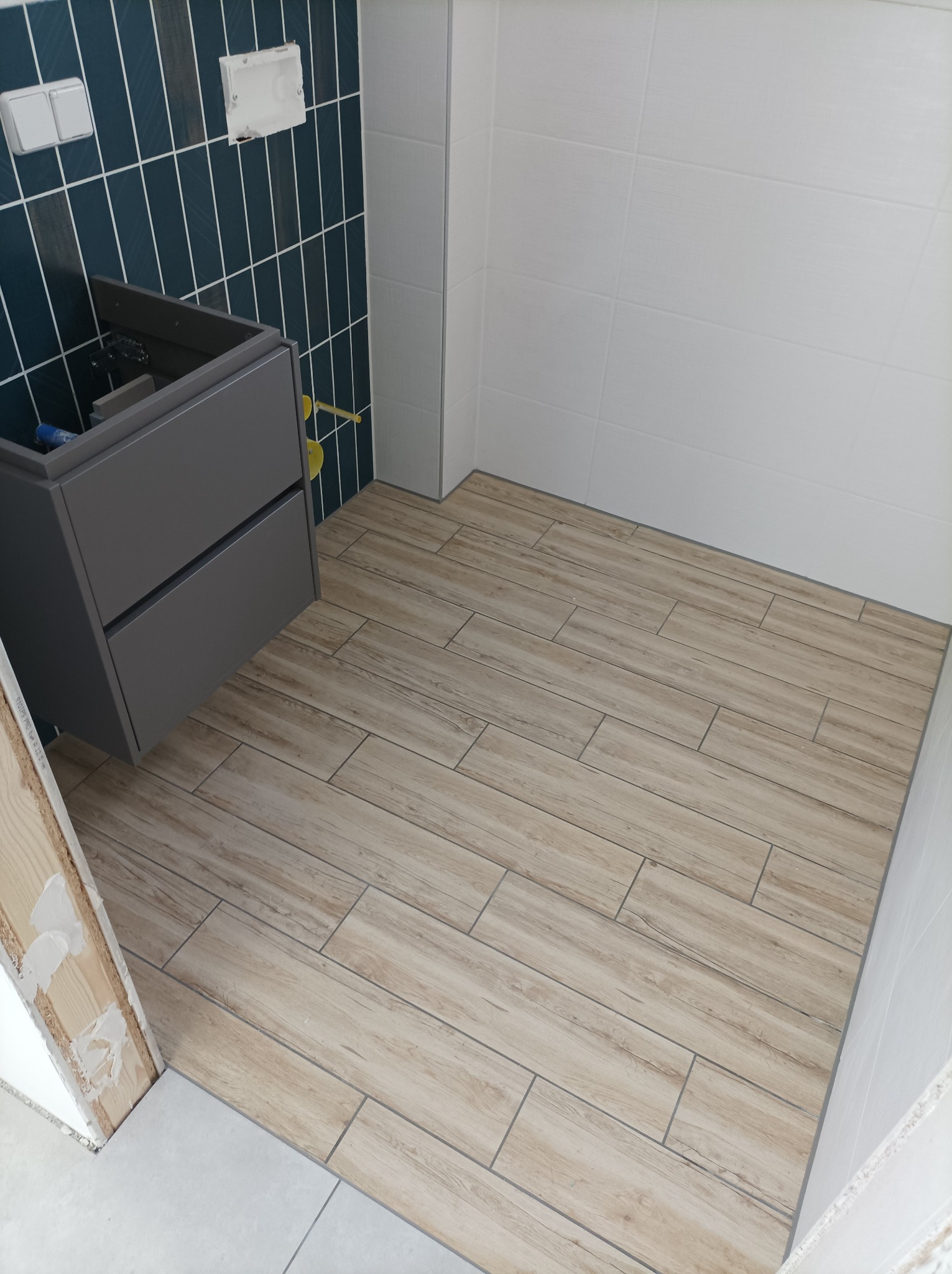 Łazienka w trakcie wykańczania: drewnopodobna podłoga z płytek, niebieskie płytki na ścianie, szara szafka pod umywalkę bez blatu, białe płytki na ścianie prysznica.