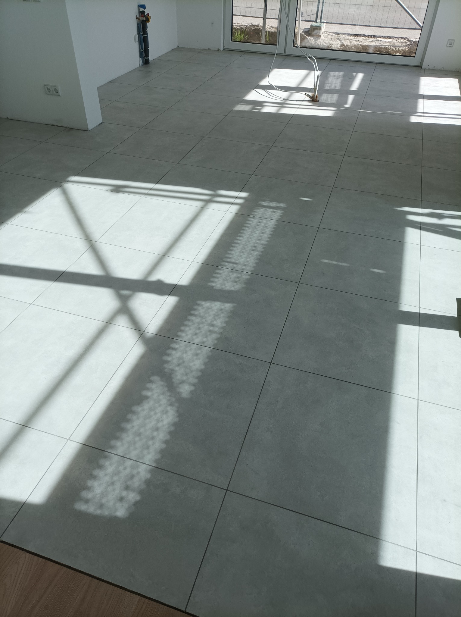 Ułożone szare płytki podłogowe w jasnym pomieszczeniu, z wyraźnym cieniem rzucanym przez okno, widoczne instalacje przy ścianie i fragment drewnianej podłogi przy krawędzi kadru.