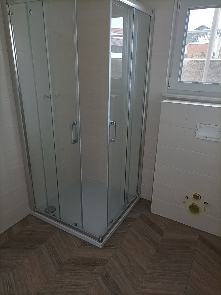 Nowa kabina prysznicowa z przesuwanymi drzwiami w narożniku łazienki wykończonej białą płytką i parkietem w jodełkę.