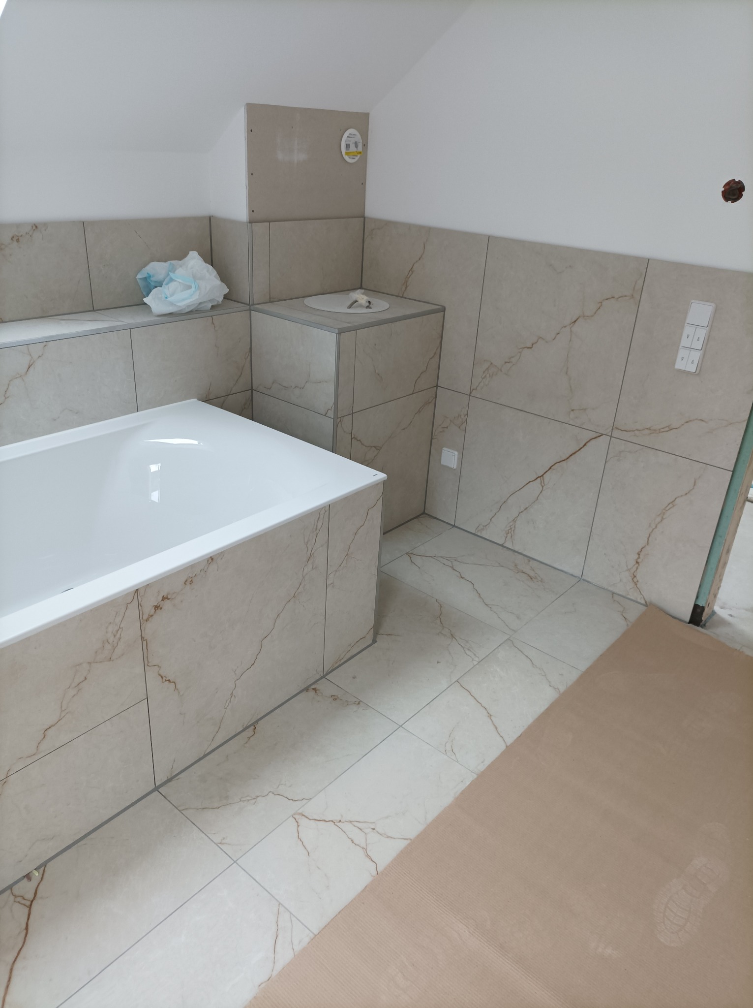 Biała wanna obudowana beżowymi płytkami imitującymi marmur, z podobnymi płytkami na ścianach i podłodze, widoczna część łazienki w trakcie wykańczania, zabezpieczona podłoga.