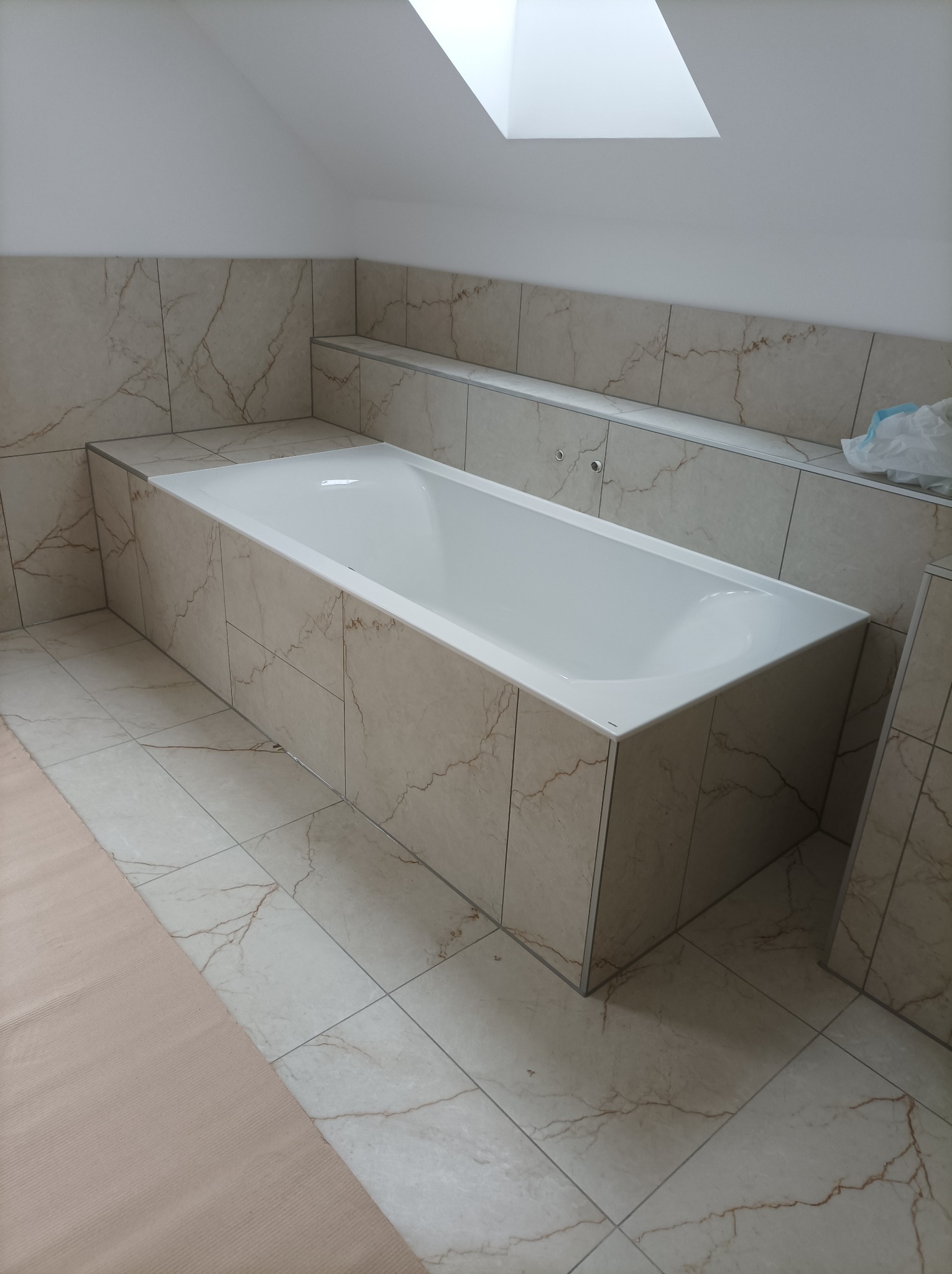 Wykończona łazienka na poddaszu z wanną obudowaną płytkami imitującymi marmur, widoczne skosy sufitowe i okno dachowe.