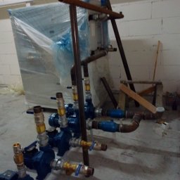 OWN Energy - Zestaw niebieskich pomp wodnych z mosiężnymi złączkami i rurami, część instalacji zapakowana w folię, widoczne rury miedziane, w tle biała ściana z pustaków i prowizoryczny stołek.