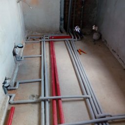 OWN Energy - Surowe wnętrze w trakcie remontu z widocznymi rurami instalacji wodnej i grzewczej w kolorach szarym, czerwonym i niebieskim, częściowo zamontowanymi na podłodze, oraz narzędzia do ich montażu.