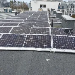 Rząd paneli fotowoltaicznych na płaskim dachu budynku mieszkalnego, widok z góry, szare niebo w tle.