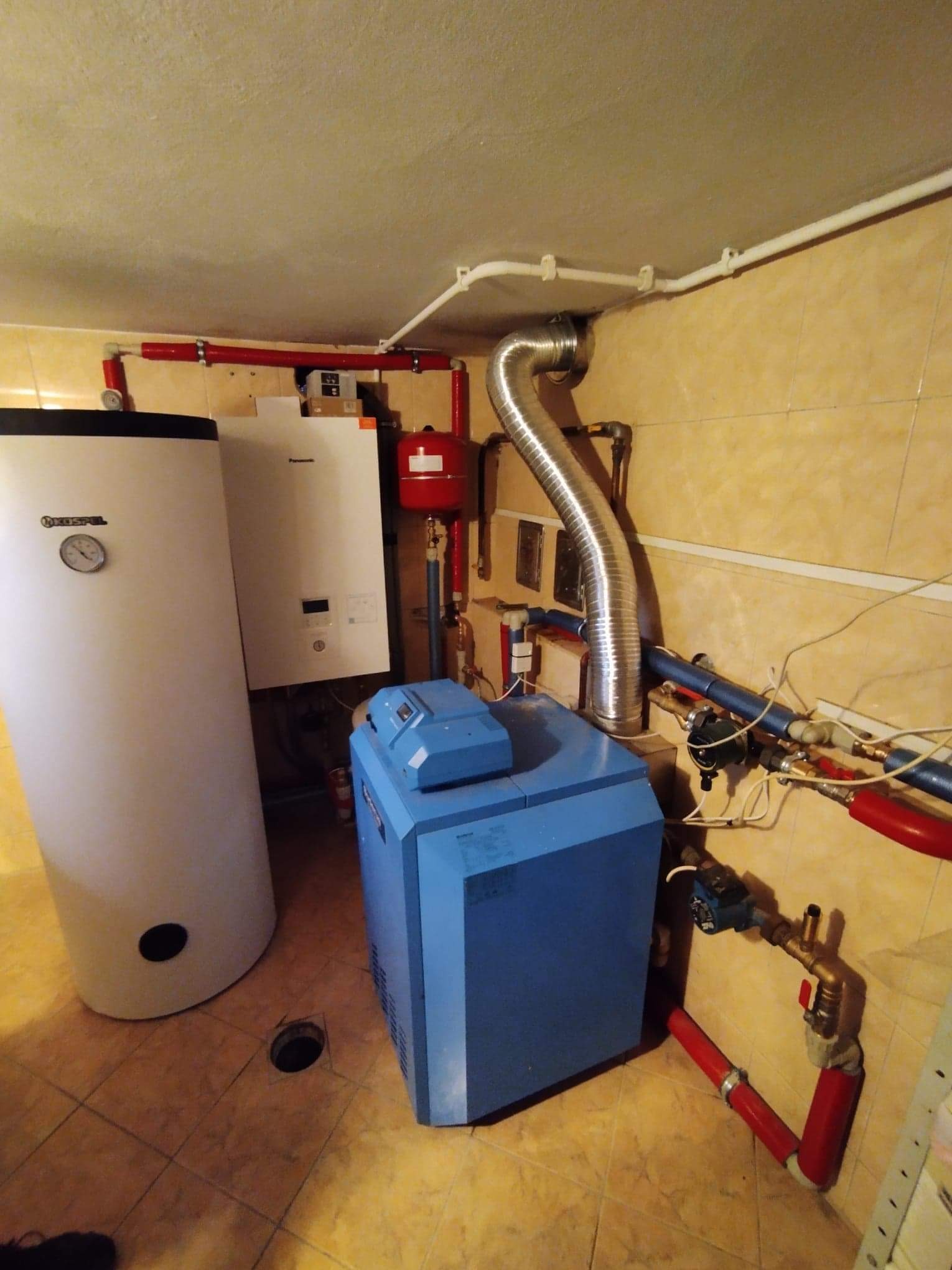 Kotłownia z piecem na paliwo stałe, zasobnikiem ciepłej wody użytkowej i kotłem gazowym, widoczne instalacje w rurach miedzianych i plastikowych, pomieszczenie wykończone płytkami ceramicznymi.