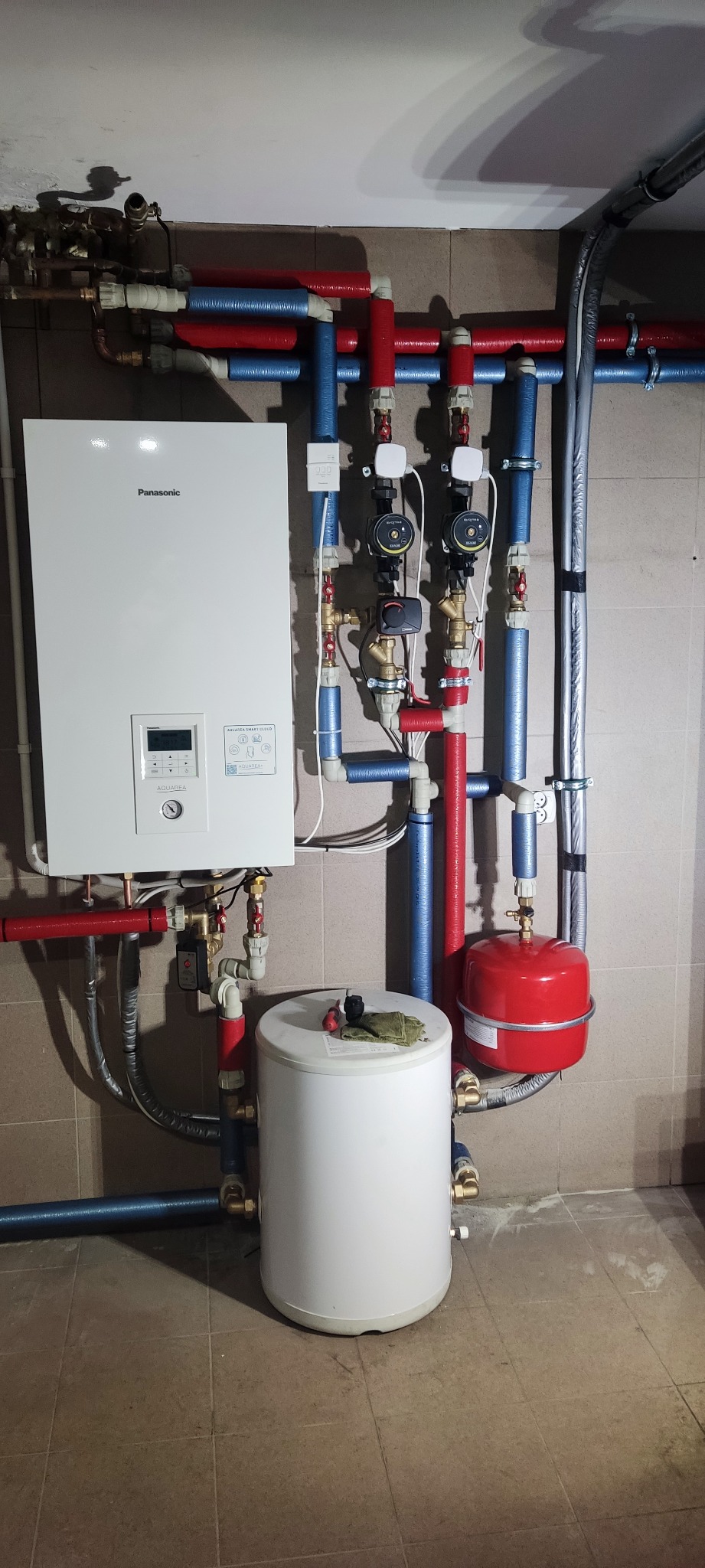 Biały piec gazowy Panasonic z wyświetlaczem LCD, pod nim biały zbiornik na wodę, czerwony zbiornik wyrównawczy, rury w izolacji czerwonej i niebieskiej, na tle płytek ceramicznych.