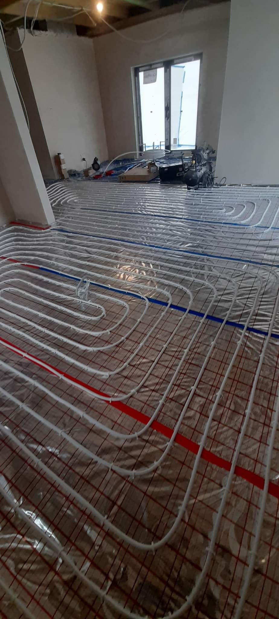 Rozłożona instalacja ogrzewania podłogowego z białych rur na folii izolacyjnej z czerwoną i niebieską taśmą, widoczne narzędzia i elementy instalacji w tle, surowy stan wykończenia wnętrza.