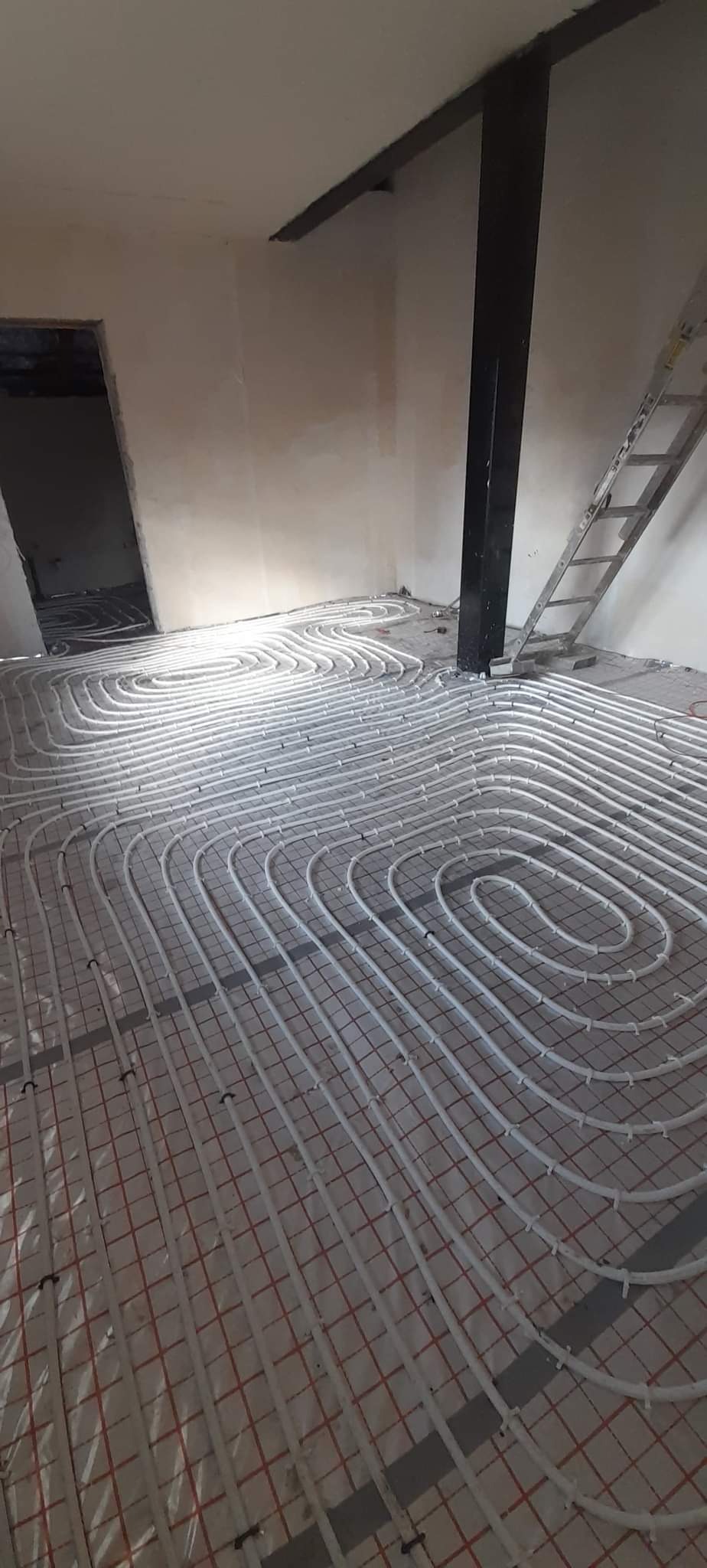 Białe rury ogrzewania podłogowego ułożone spiralnie na czerwonej siatce w surowym wnętrzu z widoczną drabiną i czarną kolumną.