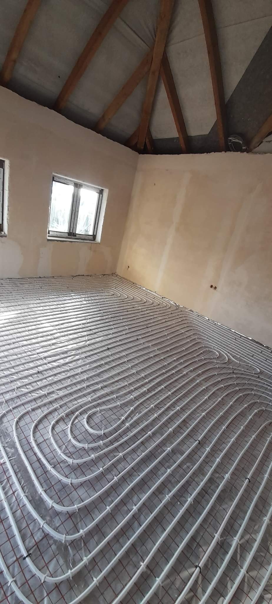 Niedokończona instalacja ogrzewania podłogowego z białymi rurkami ułożonymi na siatce w surowym pomieszczeniu z oknami i drewnianą konstrukcją dachu.