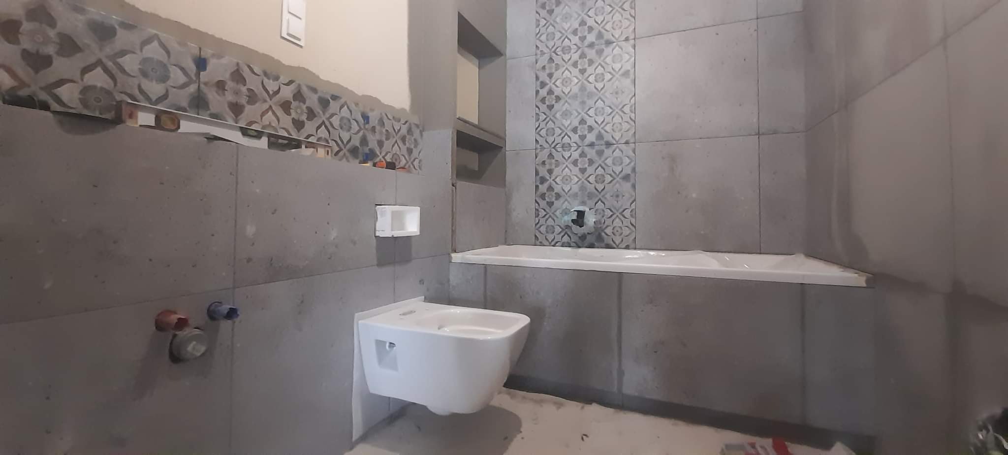 Montaż wiszącej miski WC w łazience z szarymi płytkami i dekoracyjnym pasem płytek na ścianie, widoczne przyłącza wodne i częściowo ułożone płytki.