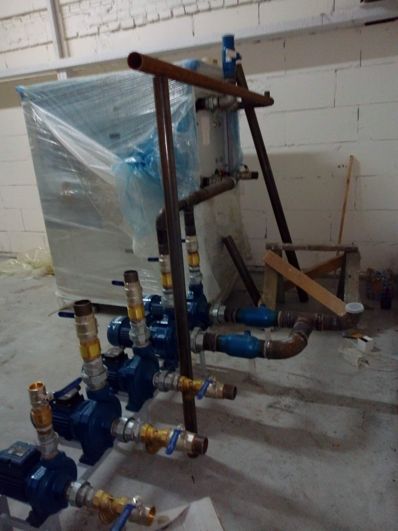 Zestaw niebieskich pomp wodnych z mosiężnymi złączkami i rurami, część instalacji zapakowana w folię, widoczne rury miedziane, w tle biała ściana z pustaków i prowizoryczny stołek.