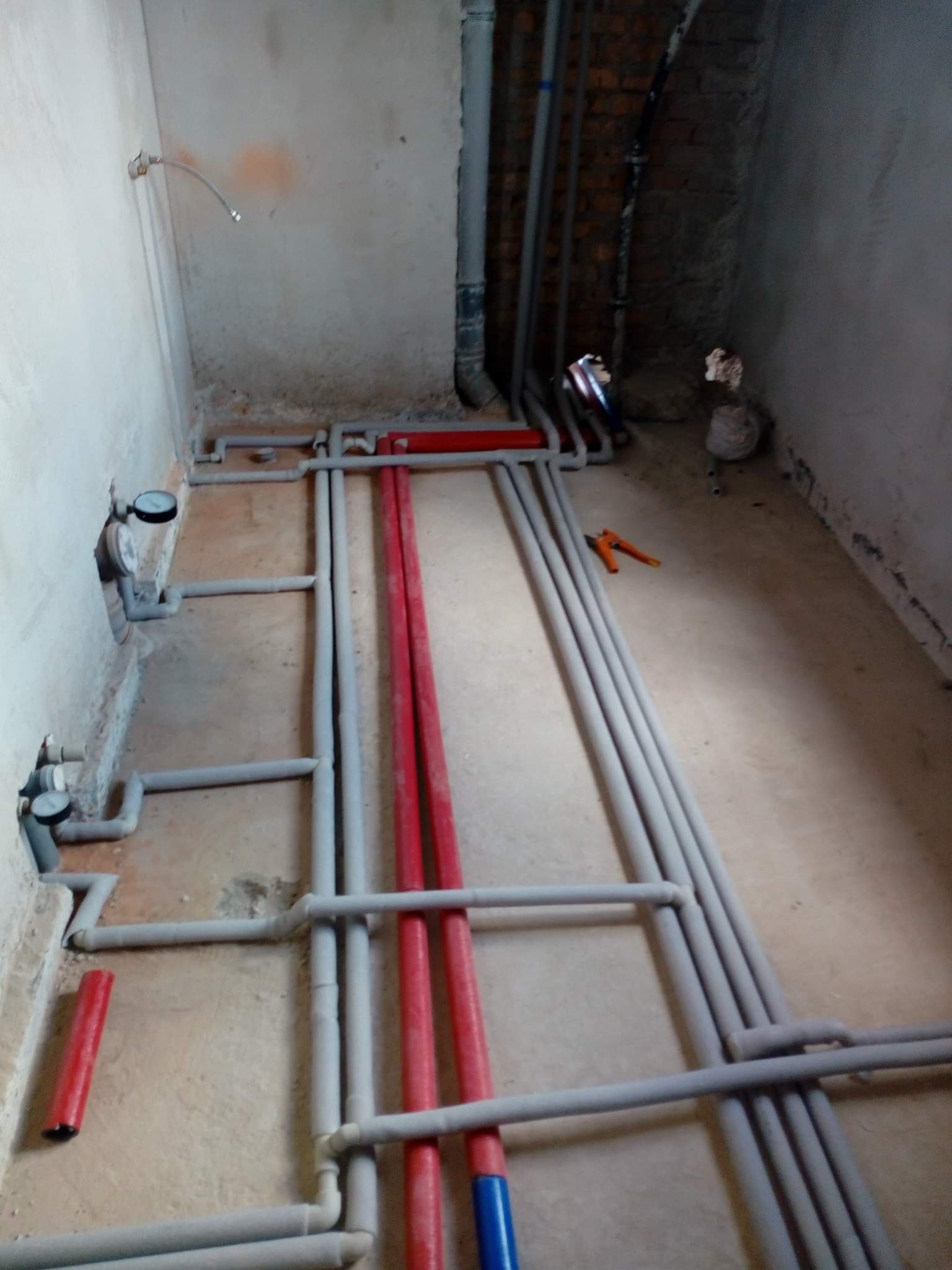 Surowe wnętrze w trakcie remontu z widocznymi rurami instalacji wodnej i grzewczej w kolorach szarym, czerwonym i niebieskim, częściowo zamontowanymi na podłodze, oraz narzędzia do ich montażu.