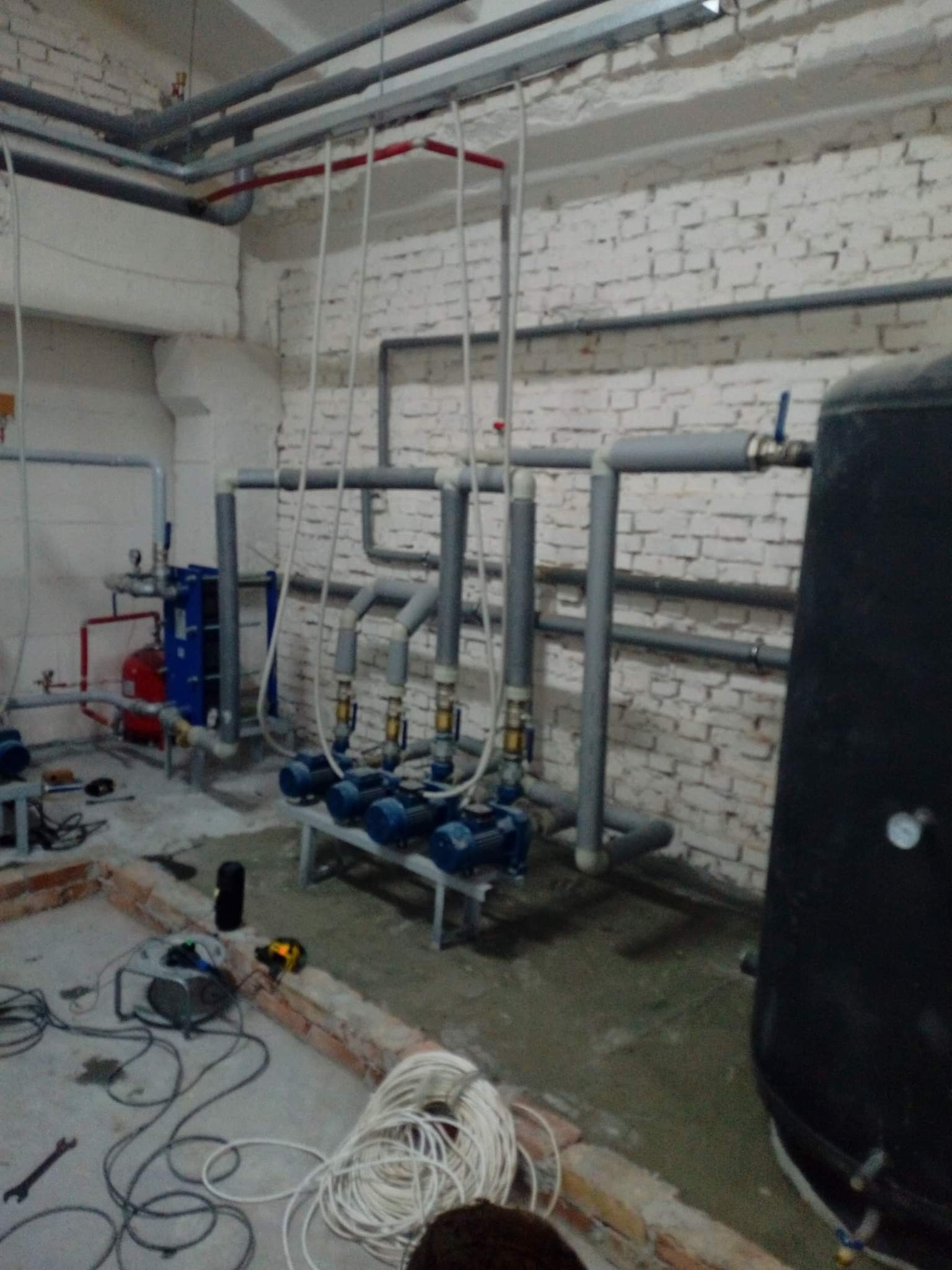 Piwnica z instalacją grzewczą: rzędy niebieskich pomp na metalowej konstrukcji, liczne szare rury pod sufitem i wzdłuż ściany z białej cegły, czerwony zbiornik, przewody elektryczne i narzędzia...