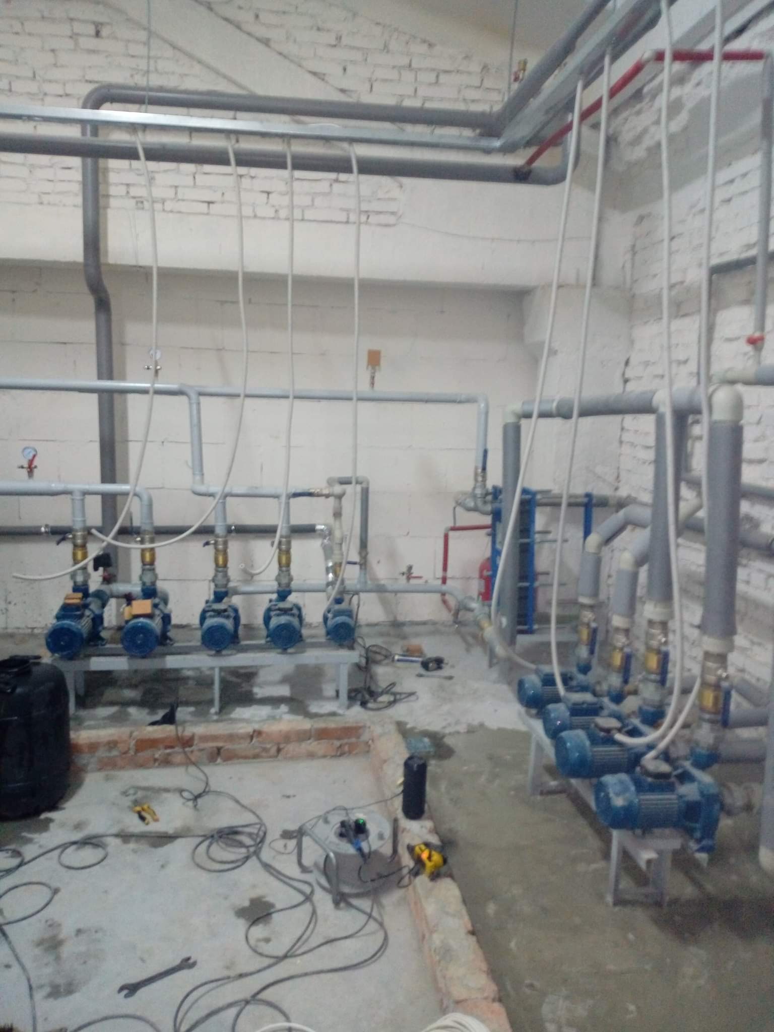 Piwnica z widocznymi niebieskimi pompami wodnymi na metalowych stelażach, szare rury instalacji wodnej pod sufitem z białej cegły, narzędzia na podłodze w trakcie montażu.