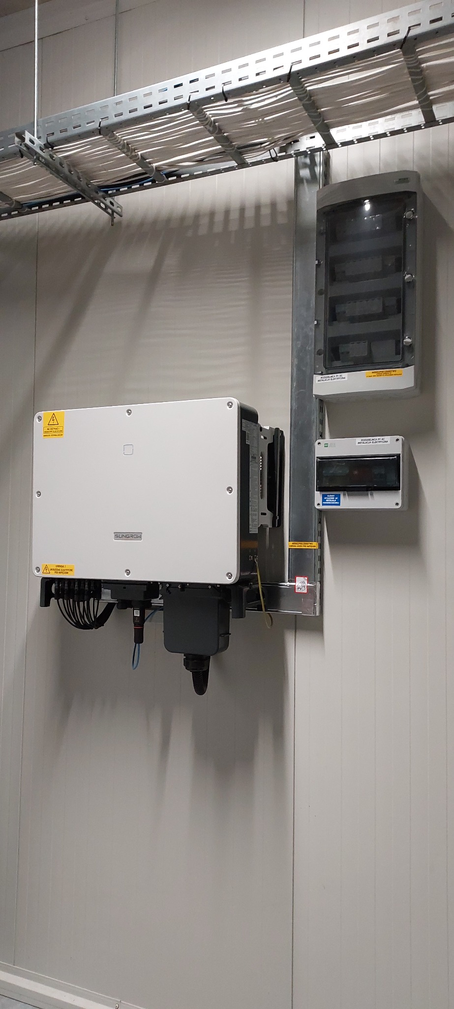 Biały inwerter solarny marki Sungrow zamontowany na ścianie z widocznymi przewodami i skrzynkami elektrycznymi, instalacja w pomieszczeniu o panelowych ścianach.