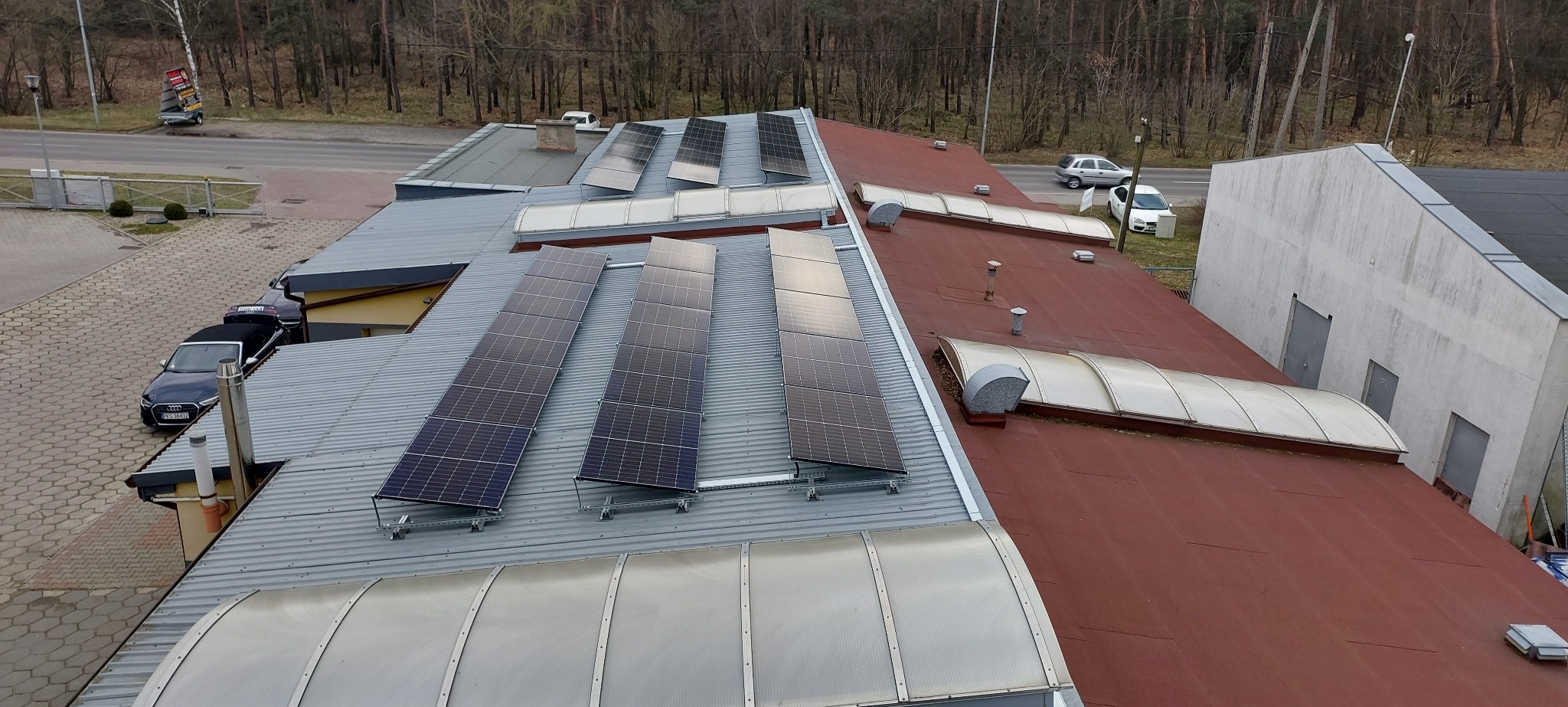 Widok z góry na zainstalowane panele słoneczne na dachu budynku z blachy trapezowej, z widocznymi świetlikami dachowymi i elementami wentylacji, w tle drzewa i droga.