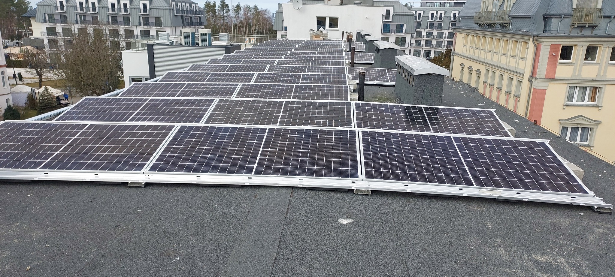 Rząd paneli fotowoltaicznych na płaskim dachu budynku mieszkalnego, widok z góry, szare niebo w tle.