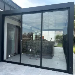 Nowoczesna, przeszklona pergola z czarnymi ramami na tarasie z szarych płyt. Widoczny fragment ogrodu i drabina wewnątrz konstrukcji, podkreślająca przestronność.