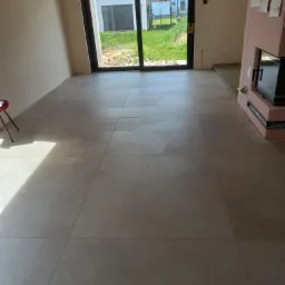 Beżowe płytki podłogowe ułożone w nowym domu, widok na przeszklone drzwi tarasowe i ogród. Fragment kominka z obudową. Minimalistyczny design wnętrza.