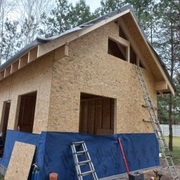 BCS - Budowa domu w trakcie realizacji: szkielet z płyt OSB, okna bez szyb, drabiny, narzędzia budowlane. Widoczne drzewa w tle.