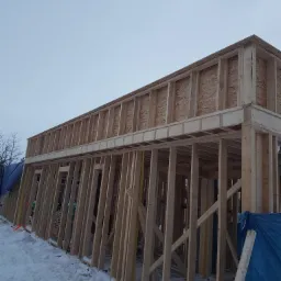 Surowa konstrukcja drewniana budynku w zimowej scenerii. Widoczne deski i płyty OSB tworzące szkielet domu, zasłonięte fragmenty plandeką, pokryte śniegiem.